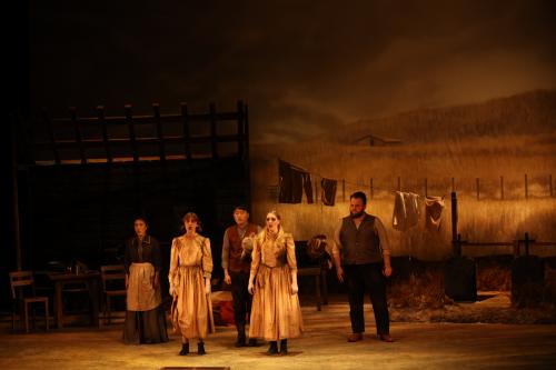 singers clad in dreary pioneer dress sing to the audience on a stage lit dimly in orange light, as if at sunset.