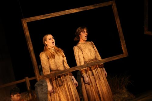 two sickly looking young women stare through a crooked window frame, singing with creepy, forlorn faces.