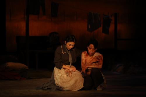 a woman crouches beside a young, shirtless boy who looks sullen and defeated. He is smeared in blood, with bloody handprints on his torso. 