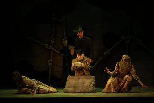 a man squats on the ground reading a letter, two women crouch near him reacting in agony to the contents of the letter. 