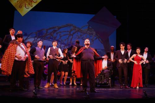 production still a group of singers on stage, in formal dress. a singer stands in front, arms outstretched, wearing a red cape. 