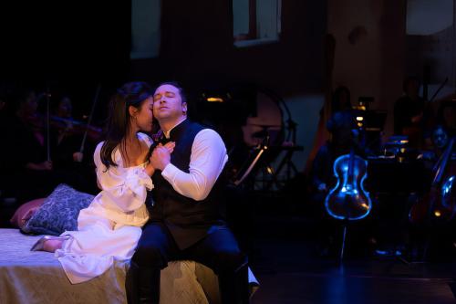 Production still from Shakespeare Gala, a man and woman hold each other closely, seated on a chaise lounge, she buries her head into his shoulder while he looks off into the distance.