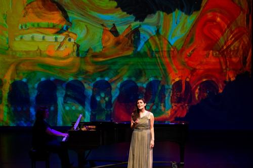 a singer in a formal gown sings on stage, accompanied by a pianist, a bold postmodern background is projected behind them.