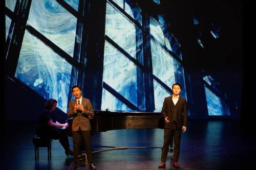 Two singers in dark suits sing on stage, behind them is a projection of a wild electromagnetic storm swirling outside of a giant wall of windows.