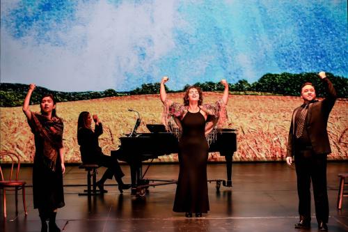 Three singers stand down stage, fists raised in the air in triumph. A Van Gogh inspired painting of a sunlit wheat field is projected behind them. 