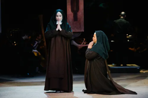 A nun stands prayerfully on stage with her head bowed, while a second nun kneels to her right, facing her, also with hands raised in prayer.