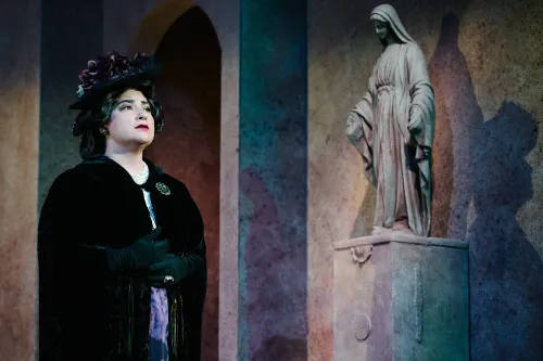 A woman stands in a dark overcoat and a fancy hat with her face toward the sky, behind her a statue of the Madonna looks down on her.