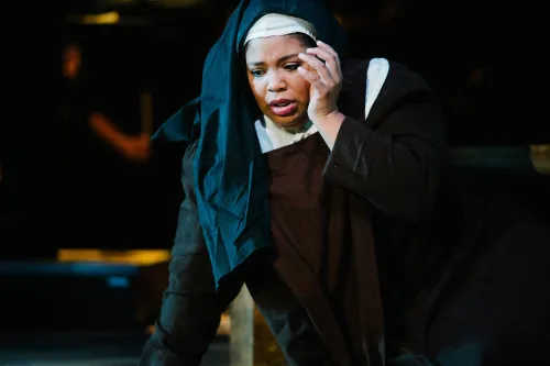 A nun sits on the ground, propping herself up with one hand and cradling her head with the other, as if she is in deep agony.