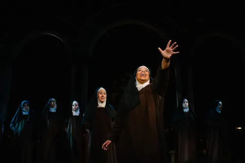 A nun stands center stage in a pool of light, with her arm stretched out in front of her, lifted hopefully to the sky, as several other nuns stand reverently behind her, fading in the darkness.