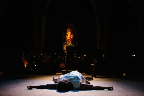 a nun in only undergarments lays splayed out on the stage floor, head toward the audience. She lays unconscious in a pool of light, the faint glow of a stained glass window behind her. The rest of the stage is in total darkness.