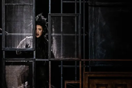 a female Ghost dressed in black stands behind the walls of a mansion, peering out at the home's inhabitants. 