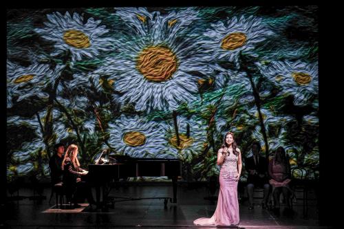 a singer in a formal dress sings in front of a concert grand piano. Behind her an expressionist painting of daisies is projected onto the background.