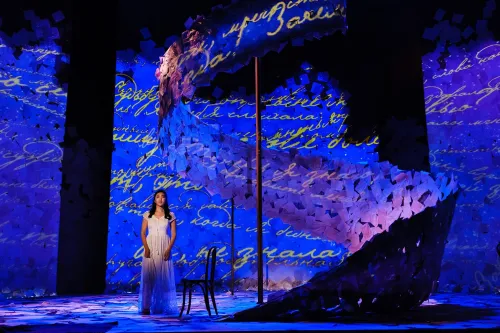 A singer in a white dress stands alone on an expansive set covered in paper, lit with lush blue light.  Cursive writing is projected all over the set. 