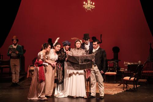 A group of people in formal Elizabethan attire stand center stage, huddled over newspaper that one woman holds up for all.