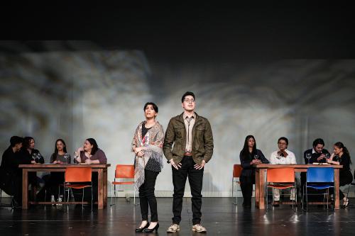 A man and woman perform together center-stage, while a chorus sits at school desks behind them.