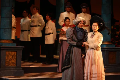 two people perform together on stage, arm in arm, while members of the chorus stand behind them.