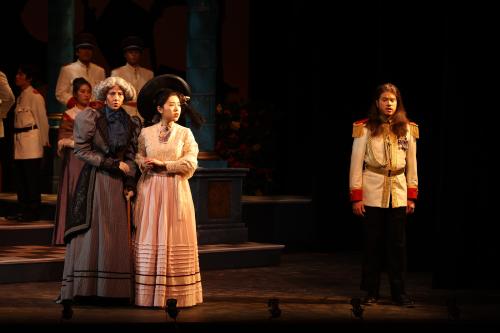 a man in military uniform stands stoically on one side of the stage, while two women stand to his right holding Hands and looking nervous.