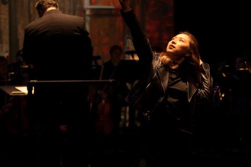 Production Still: A woman clad in black lifts a sword above her head, looking up at the blade in a powerful pose.