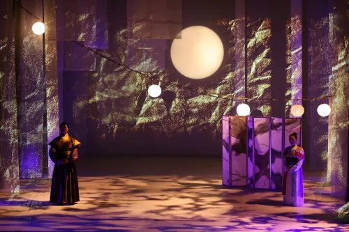 A man and woman stand on stage in traditional Chinese Dress, festoon light hang above them and a soft glowing moon is projected on stage behind them.