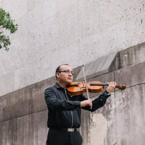 A man wearing black clothing and glasses plays the viola in front of a gray stone wall.