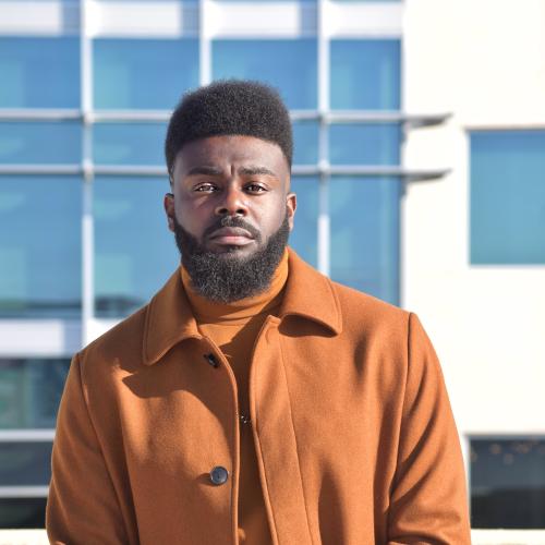 A man wearing an orange turtleneck and jacket stares seriously at the camera. He is standing in front of a glass building.