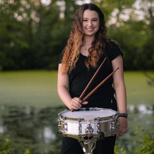 Lauren Molloy stands in an open, grassy field, behind a snare drum on a stand, and smiles into camera.