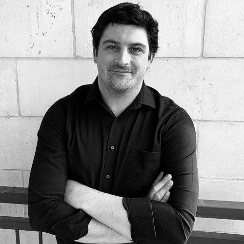 a headshot of Issa Ali, standing, arms folded, against a brick wall and smiling.