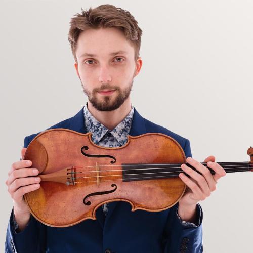 Willhelm Magner Looks into camera, holding his violin on its side.