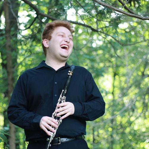 Matt Rockwell stands in a wooded area, holding his clarinet and laughing. 