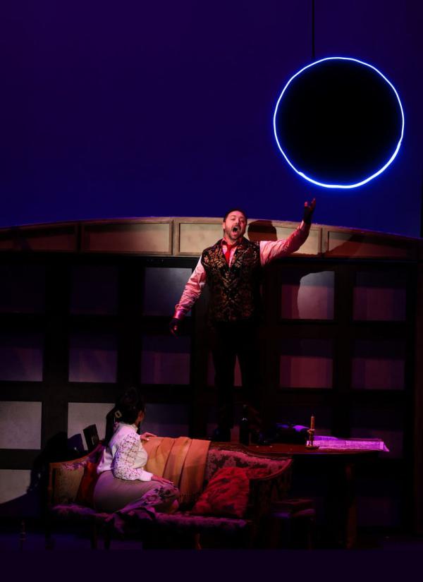 A man stands on stage with arms outstretched below a black moon with a ring of blue neon light around it. Another man sleeps on a mattress on the at his feet. 