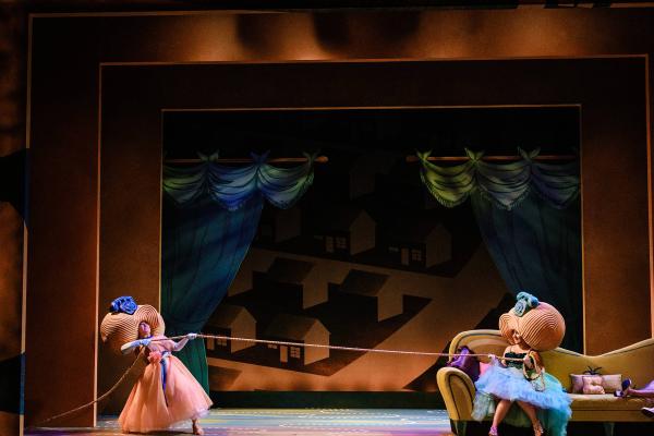 Two women stand on a cartoonish set in front of an oversized couch. Both women have huge foam beehive wigs on their heads with rotary telephones perched on top. They stand at opposite ends of the colorful set playing tug of war with a telephone cord. 