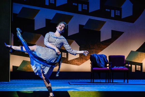 A woman in a plaid mid-century dress kicks her leg into the air as she waves a scarf around during a dance number.