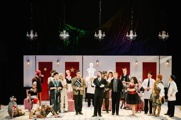 The cast of Ariadne stand on stage listening to a man in a tuxedo and white gloves as he sings.