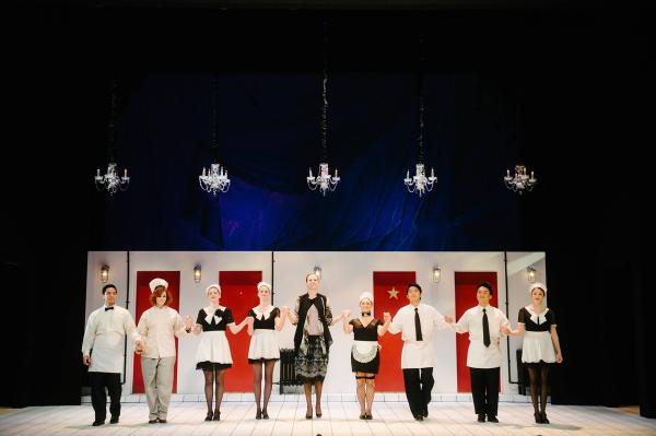 A group of maids and butlers stand in a single row on stage, hand in hand, smiling.