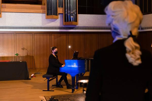 In the foreground, the back of the opera conductor can be seen, wearing a Mozart wig as she conducts, in the background is a pianist sitting at a piano on the concert hall stage.