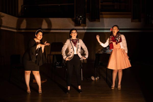 Three singers perform on a concert hall stage, dressed in modern formal wear.