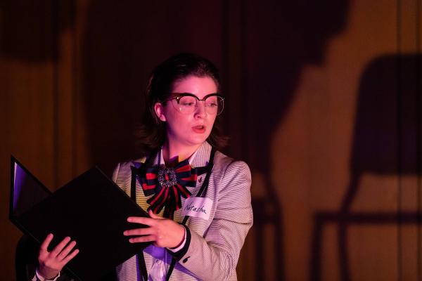 A singer on a darkly lit concert stage holds a music score in her hand, she looks down at the ground while she 'rehearses' for an upcoming audition.