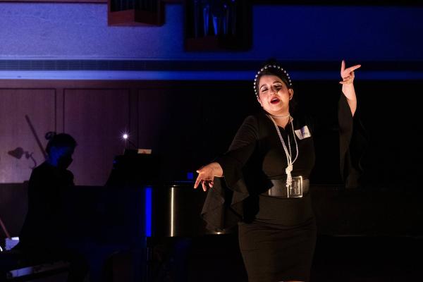 a singer in a black formal dress and pearl necklace sings exuberantly on a dark stage washed in blue light.