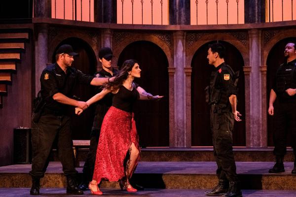 Carmen lunges at a calm and confident military officer, while two other officers hold her back.  