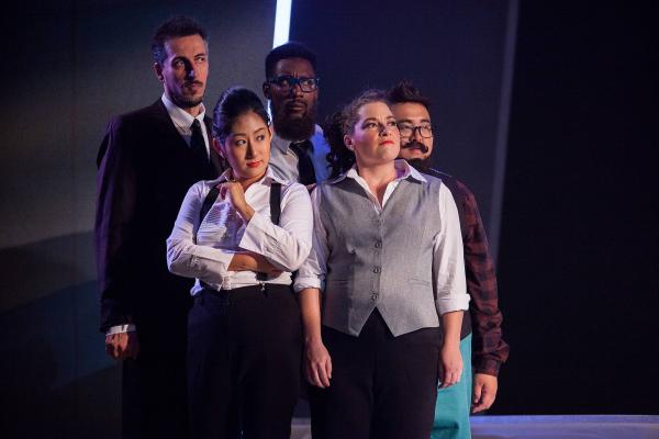 A group of singers in modern business attire huddle on center stage. 