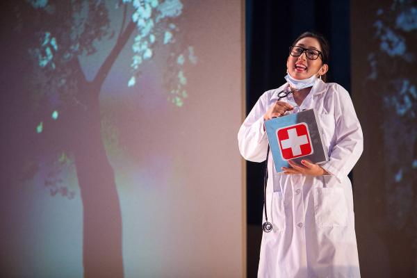 A woman stands in a medical overcoat with a stethoscope around her neck, holding a first aid kit in her hands. 