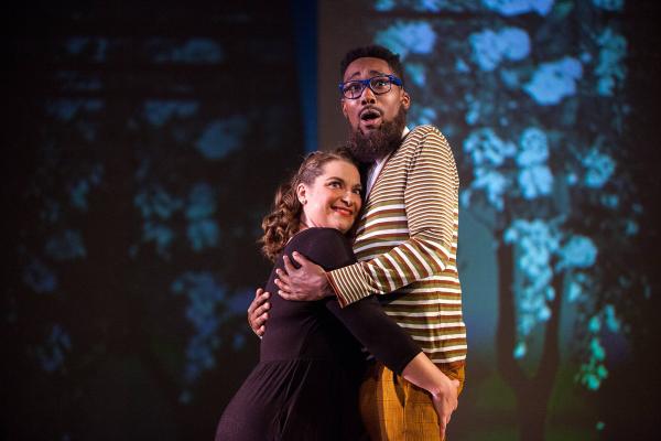 A man and woman embrace each other center stage in front of a forest projected onto the background of the stage.