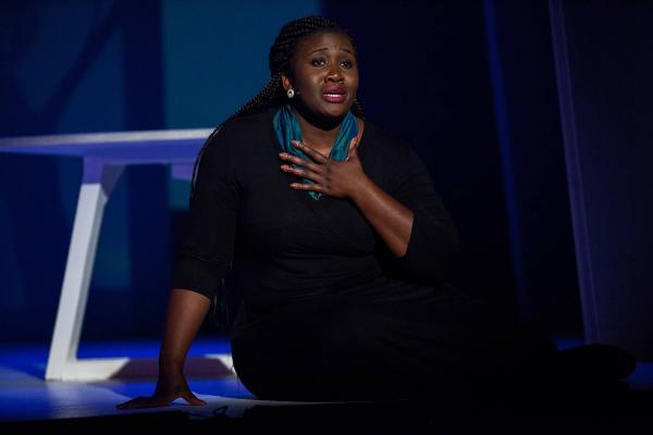 a woman sits on the ground on a very dark stage, hand gripping her chest as she sings.