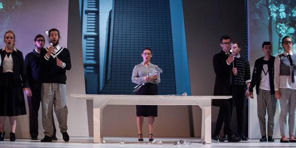 A group of singers in modern business attire stand on the set of a high-rise office building near a large white desk.