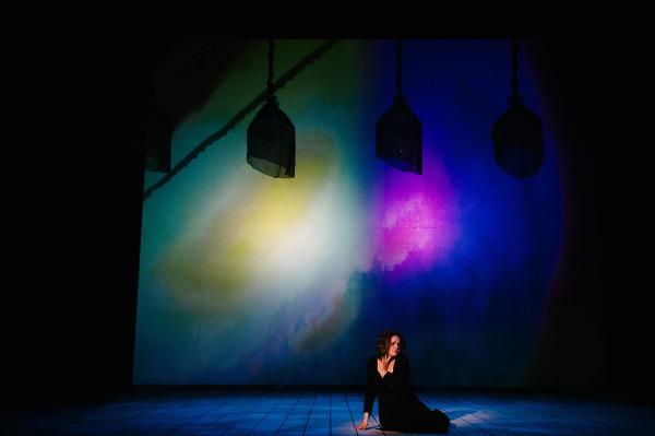 A woman sits on the stage floor, singing as she clutched her chest in anguish, behind her the stage is washed in soft blue, purple and green light.