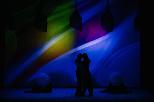 Two performers stand center stage, embracing one another, with their foreheads touching. they are lit only from behind by a deep blue light, and are surrounded by shadowy figures on both sides.