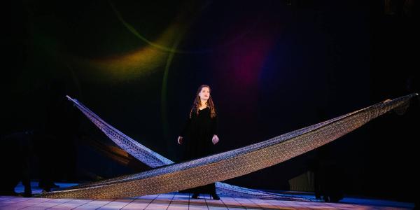 a performer in a black gown stand center stage, long strips of fabric cross-cross in front of her and behind her.