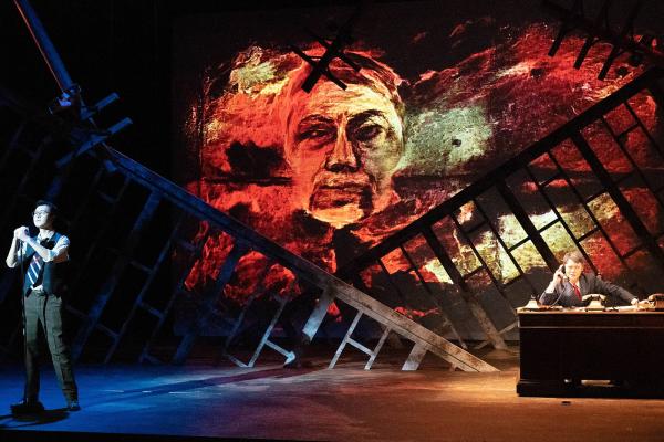 a perforer sits at a desk on one side of the stage, while another performer cowers on the other side.
