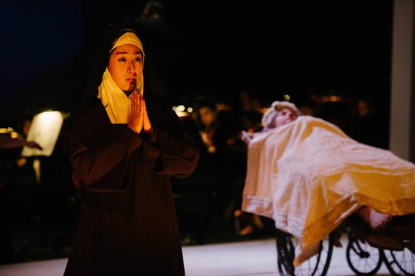 A nun kneels and prays in the foreground, a woman in a hospital bed is in the background.