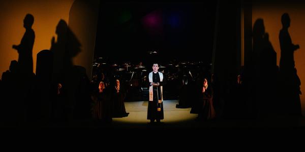 a Priest stands alone in a pool of light on a dark stage, shadows of unseen people are cast on warm orange walls in the background.